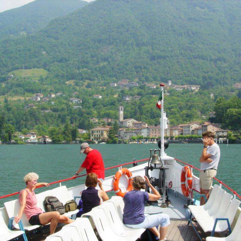 Passeggeri sul ponte di un traghetto con vista su un villaggio e colline sul Lago di Como