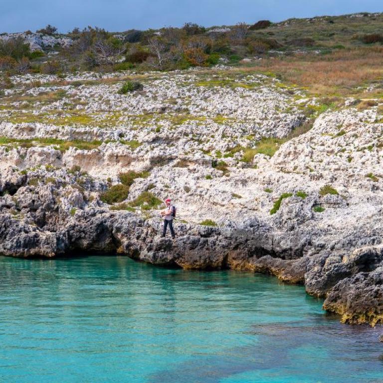 Senderistas a lo largo de la costa rocosa del Salento con mar turquesa – viajes a pie por Puglia y Matera