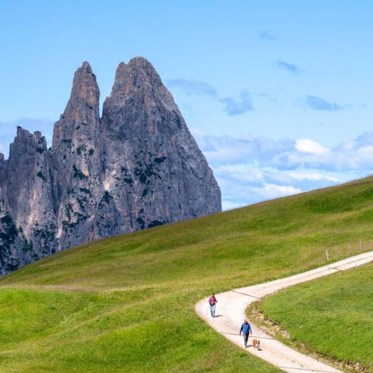 Wide view of the Dolomites from the Sentiero del Castagno, with hikers following a scenic mountain path through green fields.